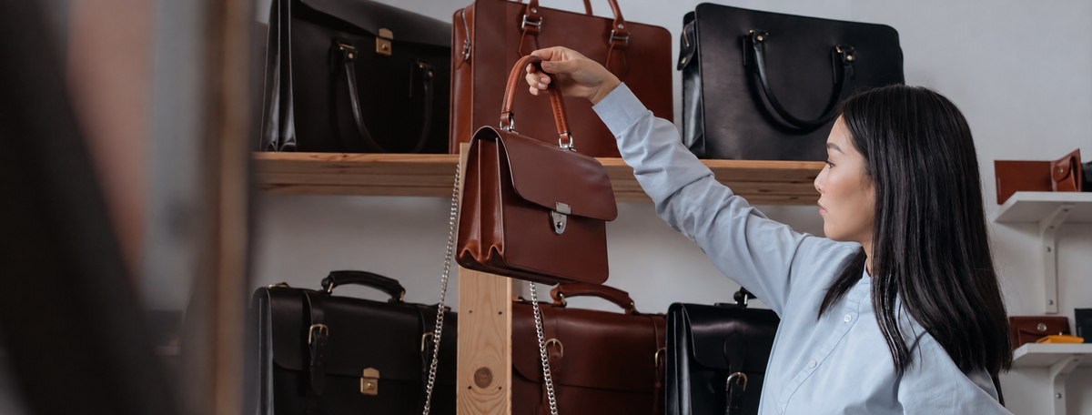 A woman in a light blue shirt reaches up to hold a brown leather handbag from a wooden display stand inside a boutique, with other leather bags on shelves in the background.